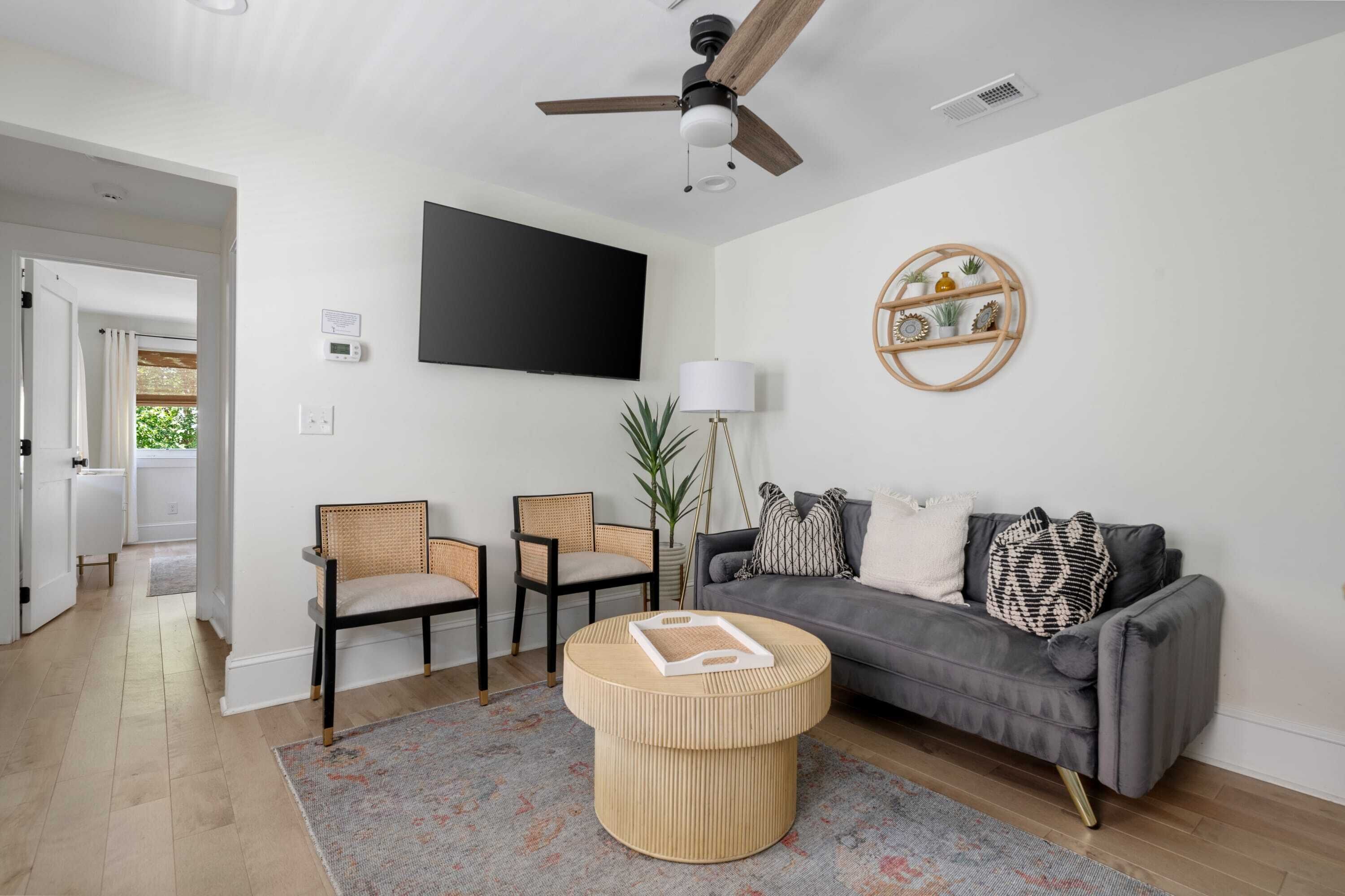 Ein modernes Wohnzimmer mit einem grauen Sofa, zwei Korbstühlen, einem runden Couchtisch, einem Deckenventilator, einem an der Wand montierten Fernseher und einem dekorativen Holzregal.