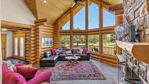 Interior de uma cabana de troncos com uma janela grande, lareira, sofá de couro preto e tapete vermelho.