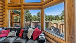 Interior de uma cabana de toras com um sofá de couro preto, várias janelas e vista para uma paisagem arborizada.