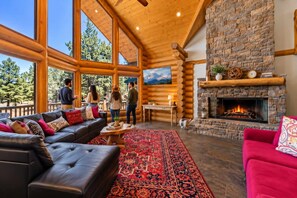 A log cabin interior with a fireplace, large windows, and a cozy seating area.