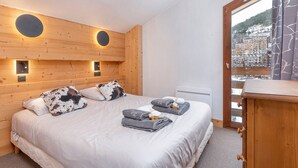 Une chambre d’hôtel avec une tête de lit en bois, deux tables de chevet, un lit avec une couette blanche et des oreillers à motifs, une fenêtre donnant sur un paysage enneigé et une porte en bois.
