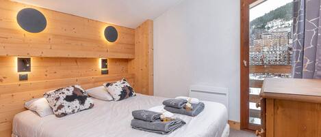 Habitación de hotel con cabecera de madera, dos mesitas de noche, cama con edredón blanco y almohadas estampadas, ventana con vistas a un paisaje nevado y una puerta de madera.