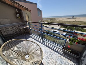 Un balcon avec un fauteuil en osier, une balustrade, des plantes en pot et une vue sur la plage et l’océan.