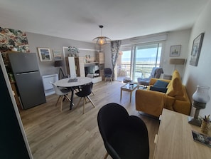 Un salon moderne avec un canapé, une table à manger et des chaises. Il y a un coin cuisine équipé d’un réfrigérateur et d’une table à manger. La pièce est dotée d’une grande fenêtre offrant une vue sur la mer.