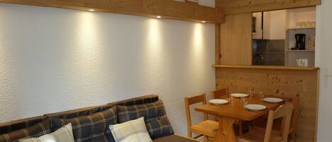 A cozy living area with a sofa, a wooden dining table, and chairs. There is a kitchen area with cabinets and a coffee maker visible.