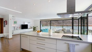 A modern kitchen with a large island, a flat-screen TV, and a view of a mountainous landscape through large windows. - Villa "Tauro 31" with Sea Views, Private Pool, and Wi-Fi (Mogán)