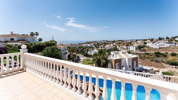 Un balcon doté d’une balustrade blanche, qui donne sur un quartier résidentiel avec des maisons et un ciel d’un bleu éclatant.