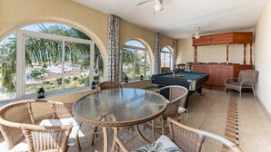 Une pièce baignée de lumière, avec une table de billard, des fauteuils en osier et une vue sur les palmiers.