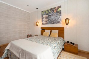 A bedroom with a bed, a headboard, a nightstand, a bedside table, a wall-mounted artwork, and two pendant lights.