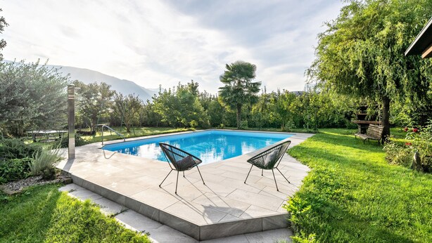 Ein rechteckiger Pool mit zwei schwarzen Liegestühlen auf einer gefliesten Terrasse, umgeben von üppiger Vegetation und einem Berg im Hintergrund.