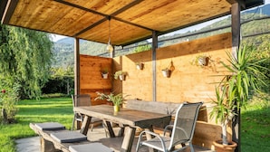 Eine Terrasse im Freien mit einem Holztisch und Stühlen, umgeben von Topfpflanzen und Blick auf eine Berglandschaft.