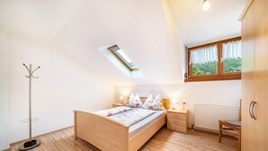 Ein Schlafzimmer mit Dachfenster, Holzboden, einem Bett mit Blumenmuster, Nachttischlampen, einem Nachttisch, einem Sessel und einem Kleiderschrank.