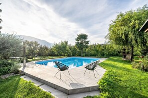 Una piscina rectangular con dos sillas negras sobre un suelo de baldosas, rodeada de exuberante vegetación y una montaña al fondo.