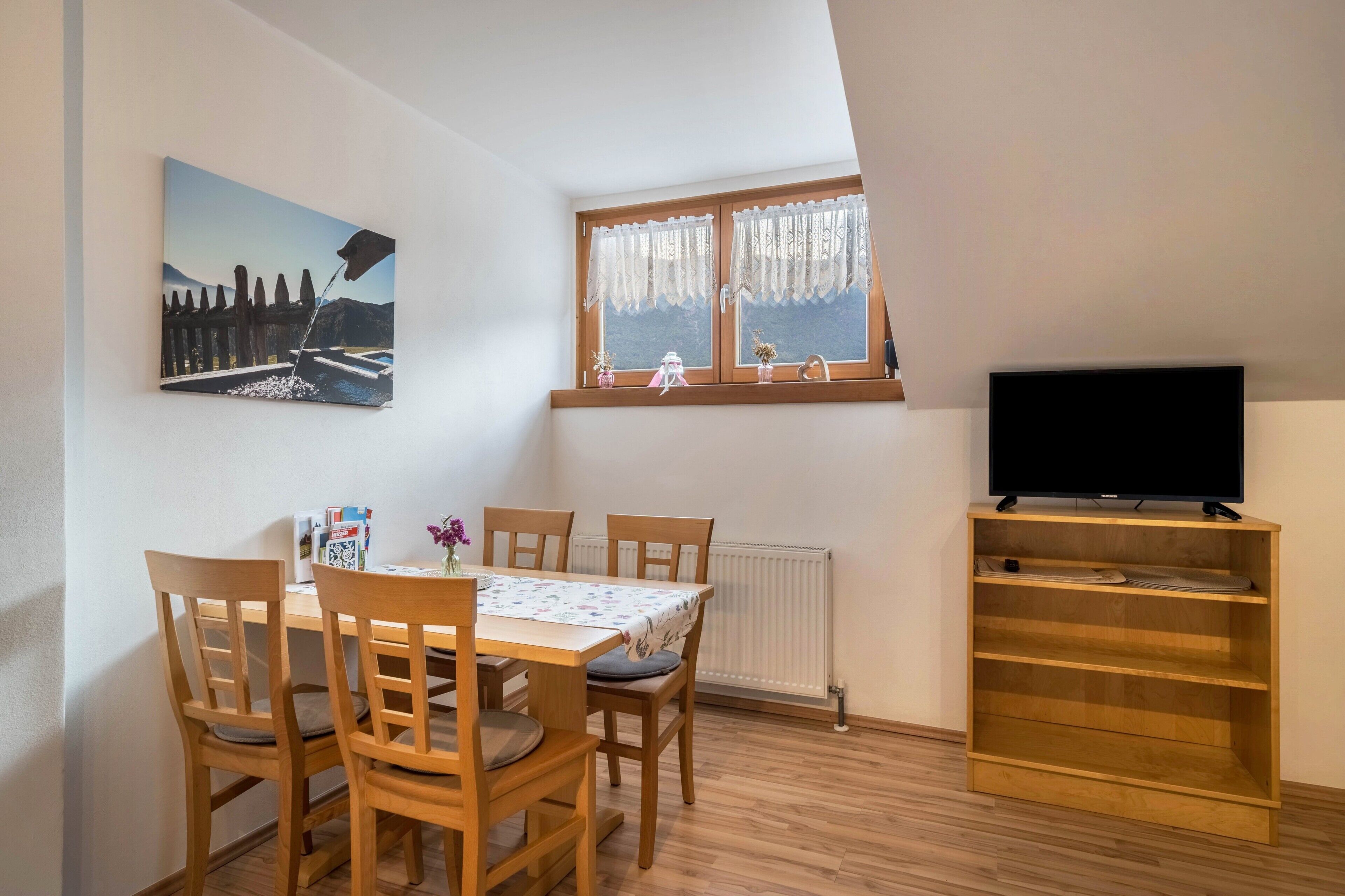 A dining area with a table and chairs, a painting on the wall, a window with curtains, and a TV on a shelf.