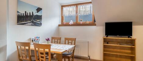 A dining area with a table and chairs, a painting on the wall, a window with curtains, and a TV on a shelf.
