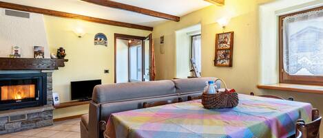 Une pièce chaleureuse avec une cheminée, un canapé, une table recouverte d’une nappe colorée et une fenêtre ornée de rideaux en dentelle.