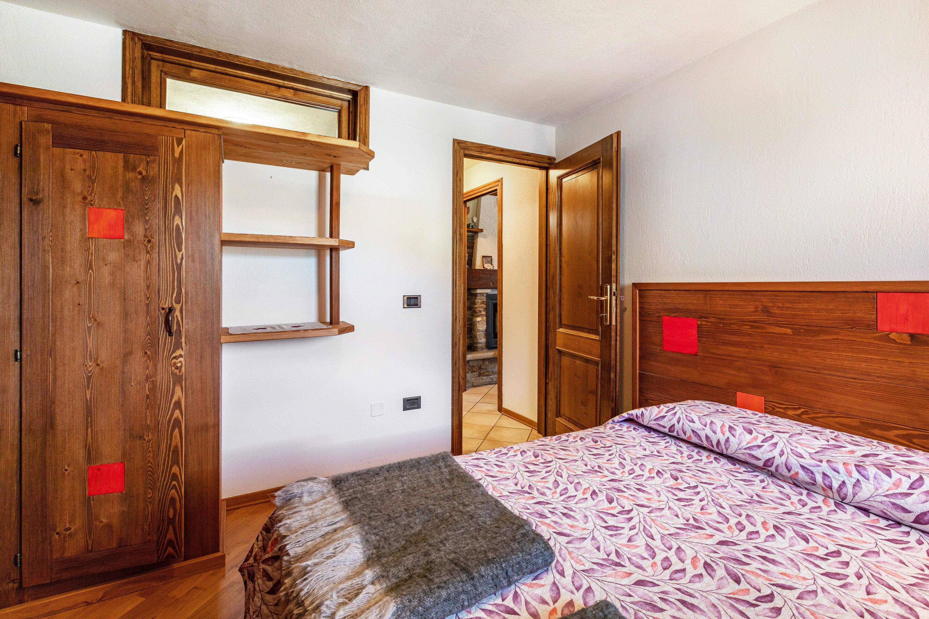 A bedroom with a wooden headboard, a bed with patterned bedding, a wooden wardrobe, and a small stool.