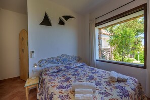 Un dormitorio con cama, mesita de noche, puerta de madera y ventana con vistas al verdor.