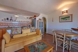 A living room with a sofa, a coffee table, a dining table with chairs, a kitchen area with a refrigerator, and a wall-mounted light fixture. - Villa 'Villetta Bomboniera' with sea view, private terrace and air conditioning (Arzachena)