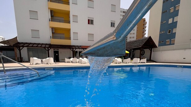 A swimming pool with clear blue water, a metal structure above it, and a multi-story building in the background. - Apartment 'Joana' with Shared Pool, Wi-Fi and Air Conditioning (Portimão)