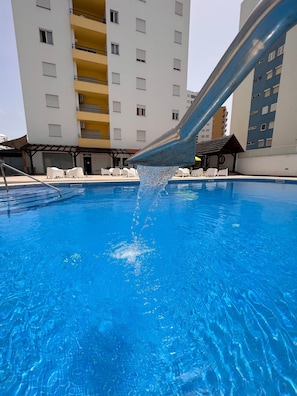 A swimming pool with clear blue water, a metal structure above it, and a multi-story building in the background.