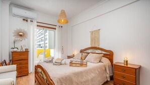A bedroom with a wooden bed, bedside tables, a hanging macrame, and a view through a window. - Apartment 'Joana' with Shared Pool, Wi-Fi and Air Conditioning (Portimão)