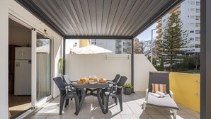 A covered patio with a table set for breakfast, outdoor chairs, a potted plant, and a view of a residential building. - Apartment 'Joana' with Shared Pool, Wi-Fi and Air Conditioning (Portimão)