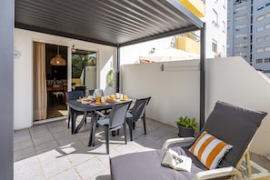 A covered outdoor dining area with a table set for two, chairs, a potted plant, and a lounge chair with a striped pillow.
