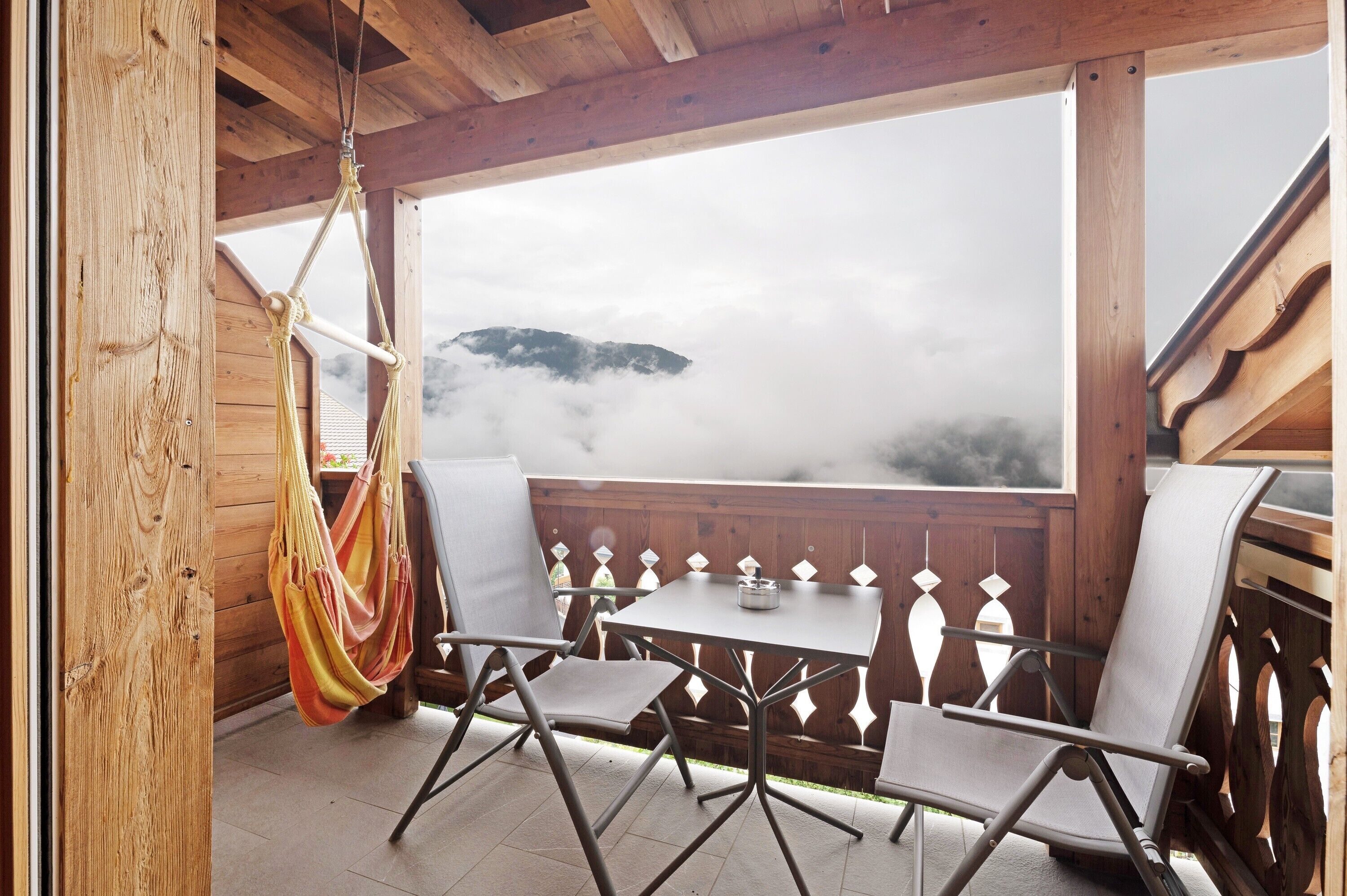 Una terraza de madera con una mesa, sillas, una hamaca y vistas a una montaña a través de una barandilla de madera.
