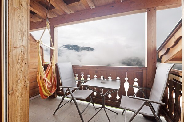 A wooden deck with a table and chairs, a hammock, and a view of a mountain through a wooden railing.