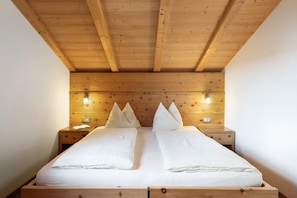 Un plafond en bois avec des poutres apparentes, un lit avec du linge de lit blanc, deux tables de chevet avec des lampes et une tête de lit en bois.