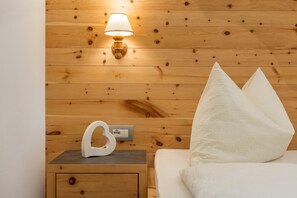 Paredes de madeira com uma luminária fixada, uma mesa de cabeceira com um objeto branco e uma cama com almofadas brancas.