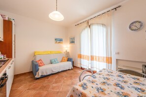 Una habitación con una cama, un sofá, una mesa con mantel estampado, un reloj de pared y un ventanal con cortinas.