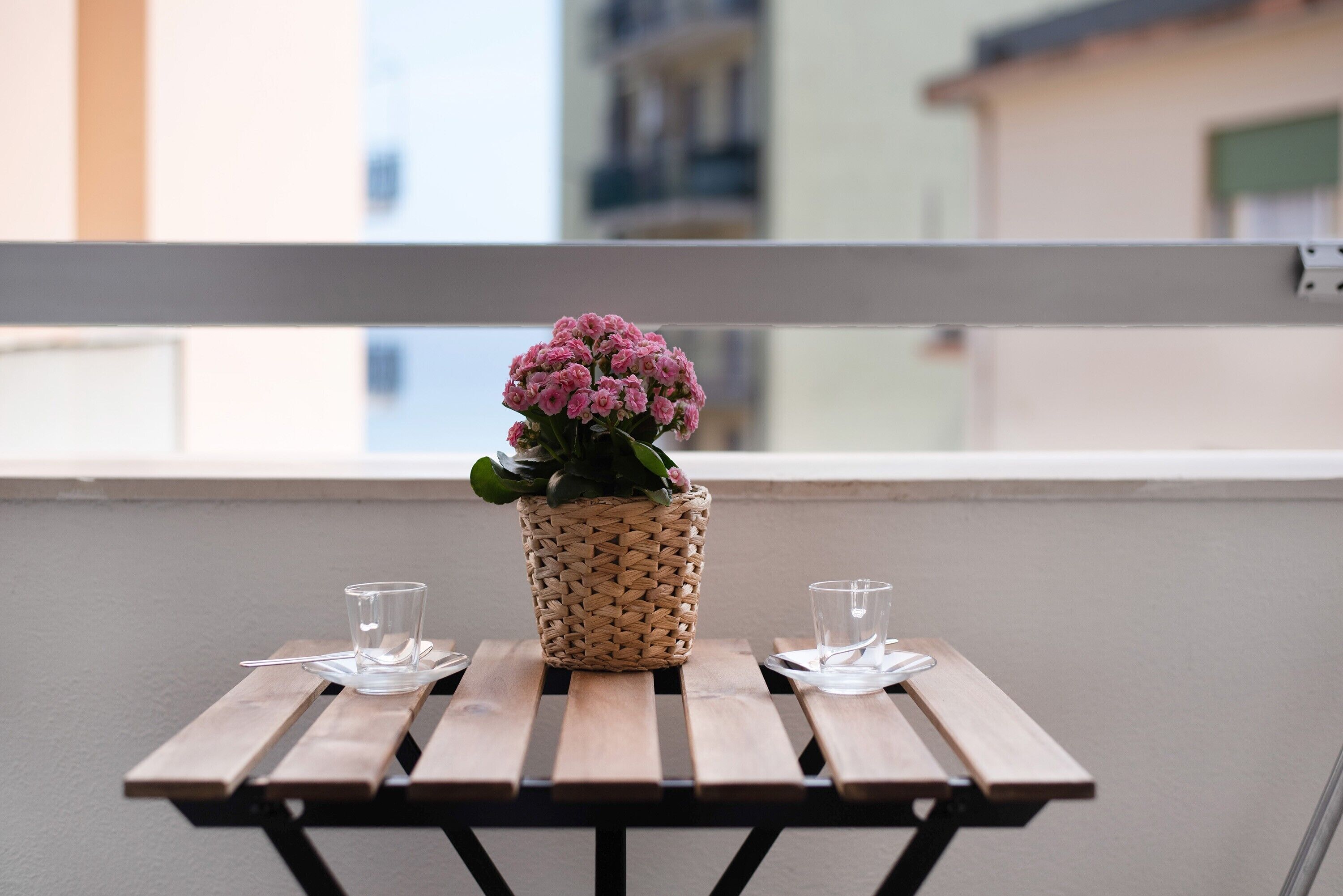 A balcony with a wooden table, two glasses, and a woven basket with pink flowers.