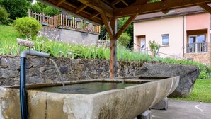 A stone water feature with a wooden structure overhead, surrounded by a stone wall and greenery. - La Sabaudia Gîte in the Vosges – Up to 10 Guests (Gérardmer)
