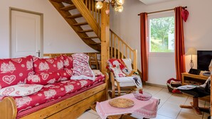 A cozy room with a wooden staircase, a red patterned sofa, a white chair, a television, and a window with curtains. - La Sabaudia Gîte in the Vosges – Up to 10 Guests (Gérardmer)