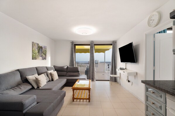 A modern living room with a grey sofa, a wooden coffee table, a flat-screen TV, and a balcony with a view.
