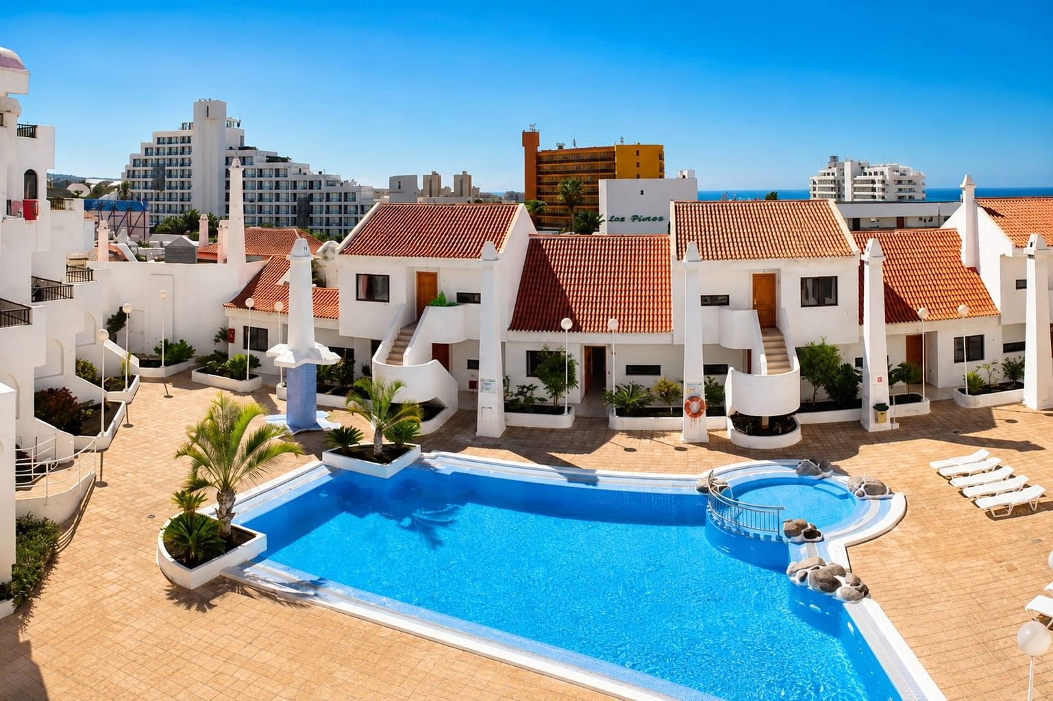 Un complejo turístico con piscina, varias construcciones con tejados rojos y vistas despejadas al mar.