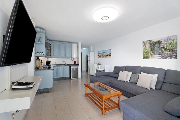 A modern living room with a grey sofa, a coffee table, a flat-screen TV, a kitchen area with light blue cabinets, and a dining table with chairs.
