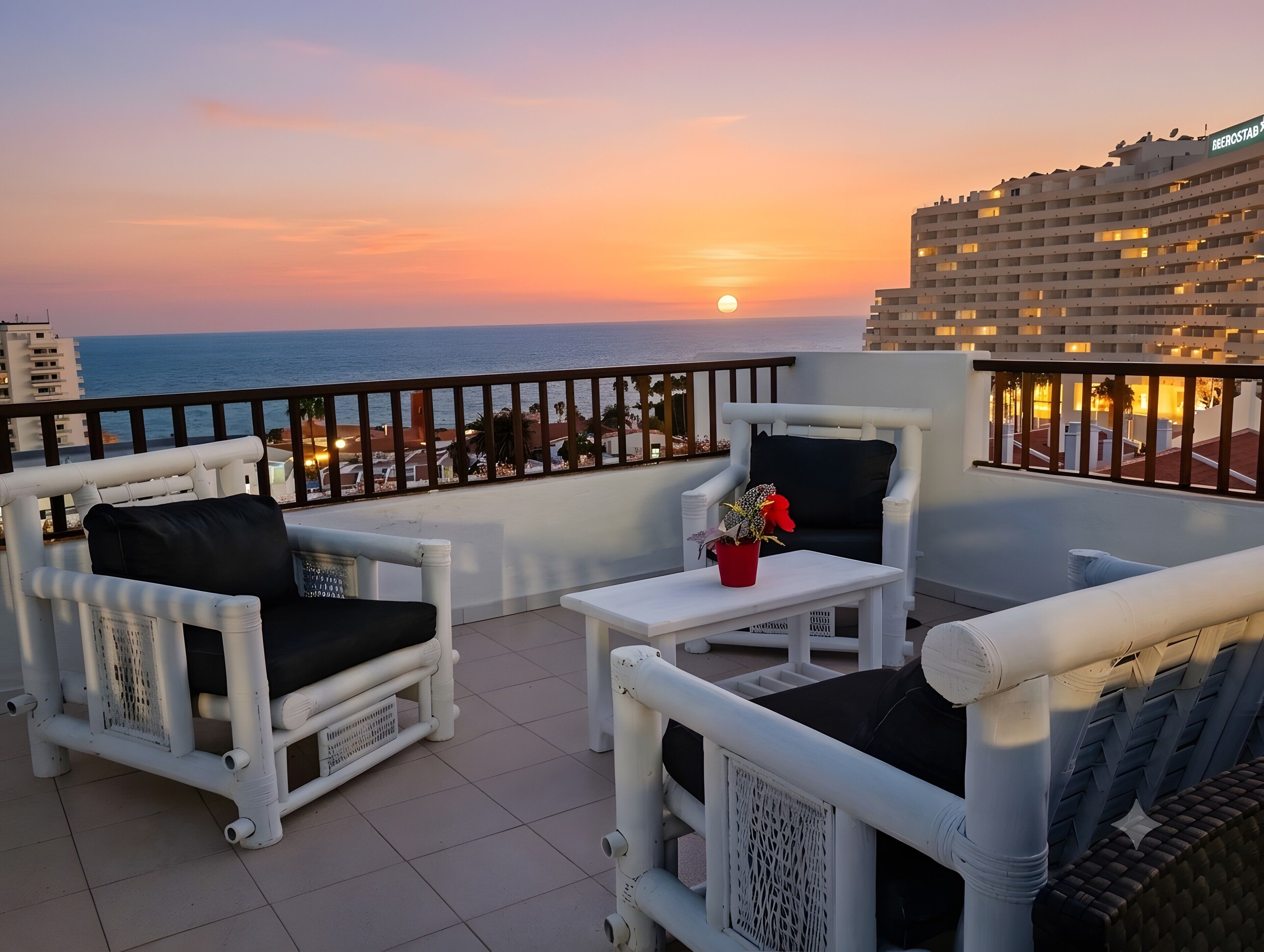 A balcony with white wicker furniture, a small table with a red pot, and a view of the sunset over the sea.