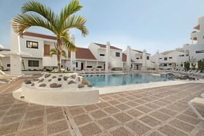 Zona de piscina con sillas de descanso, una palmera y un edificio con tejado de baldosas.