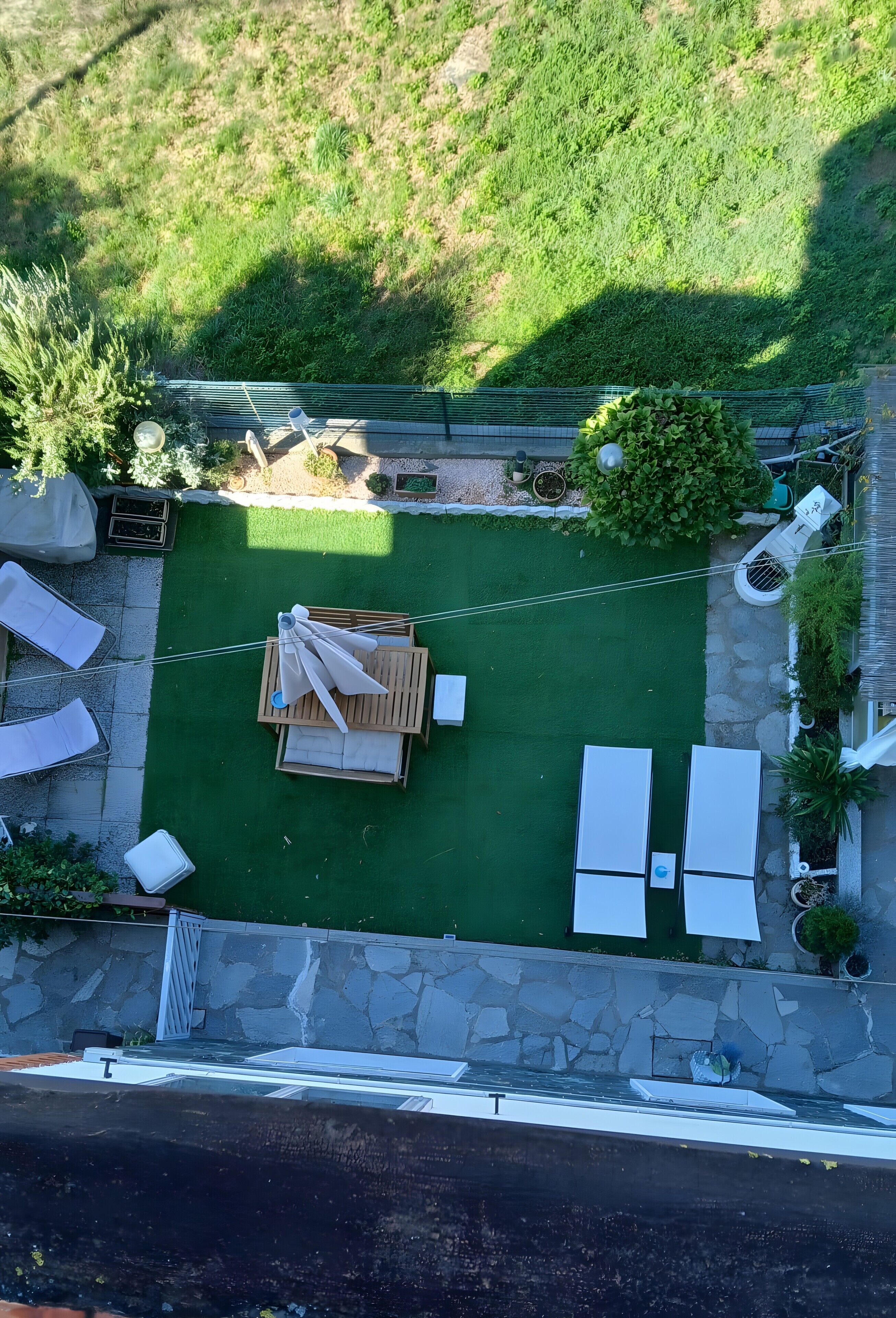 Eine Vogelperspektive zeigt einen Garten mit einem grünen Rasen, einer Holzterrasse und weißen Liegestühlen.