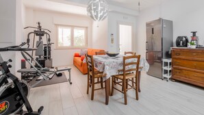 Une salle de sport équipée de matériel d’exercice, une table à manger avec des chaises, un canapé orange, un réfrigérateur et un lustre.