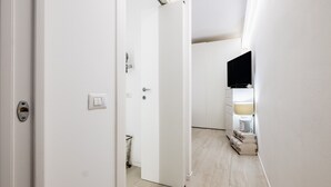 Un couloir avec une porte blanche, un interrupteur, une prise de courant, un téléviseur fixé au mur et une lampe.