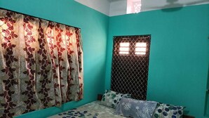 A bedroom with teal walls, a floral-patterned bedspread, and a window with a metal grille. - Puri (Puri)