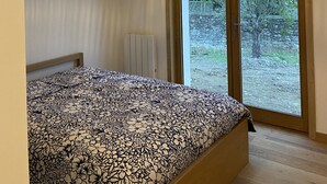 Un dormitorio con cama, lámpara colgante, ventana grande y vistas al exterior con vegetación.
