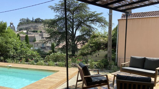 Una terraza de madera con piscina, mobiliario exterior y vista a un edificio histórico al fondo.