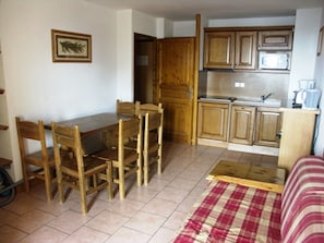 A compact kitchen with wooden cabinets, a dining table with chairs, and a bed with a plaid blanket. - Cozy 2-room apartment, parking and pets allowed, near slopes (Orcières Merlette)
