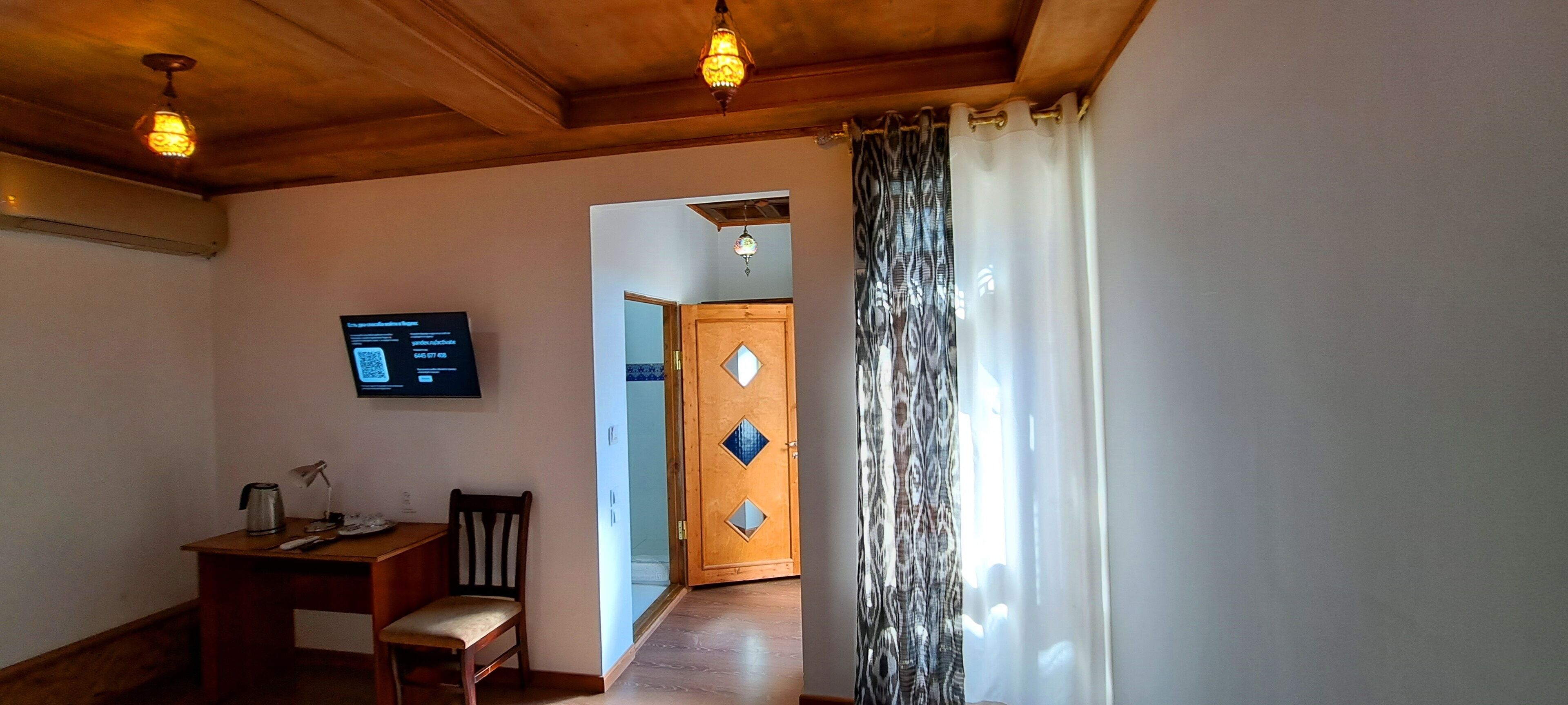 A room with a wooden ceiling, a desk with a chair, a television on the wall, and a patterned curtain.