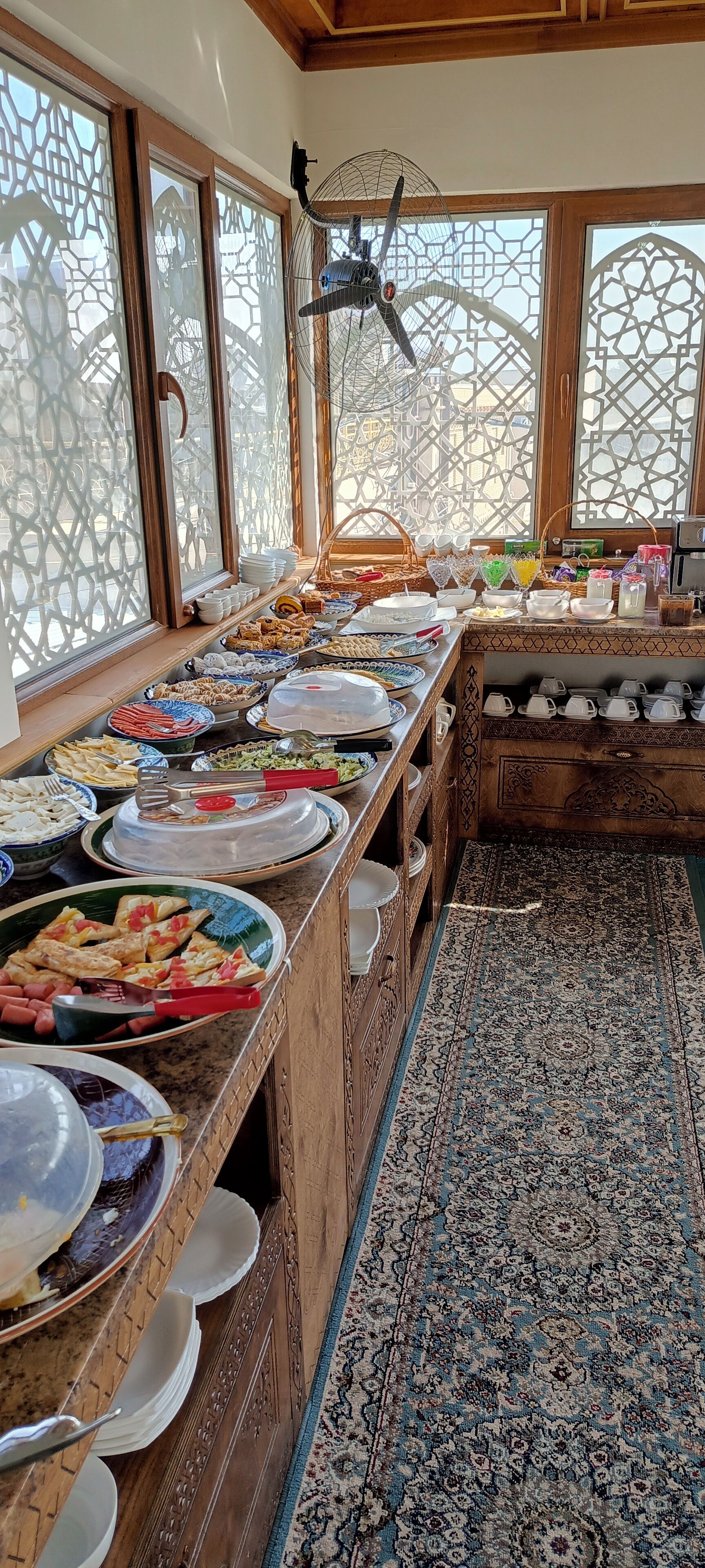 Un buffet proposant divers plats et ustensiles, un tapis à motifs et des grilles décoratives aux fenêtres.